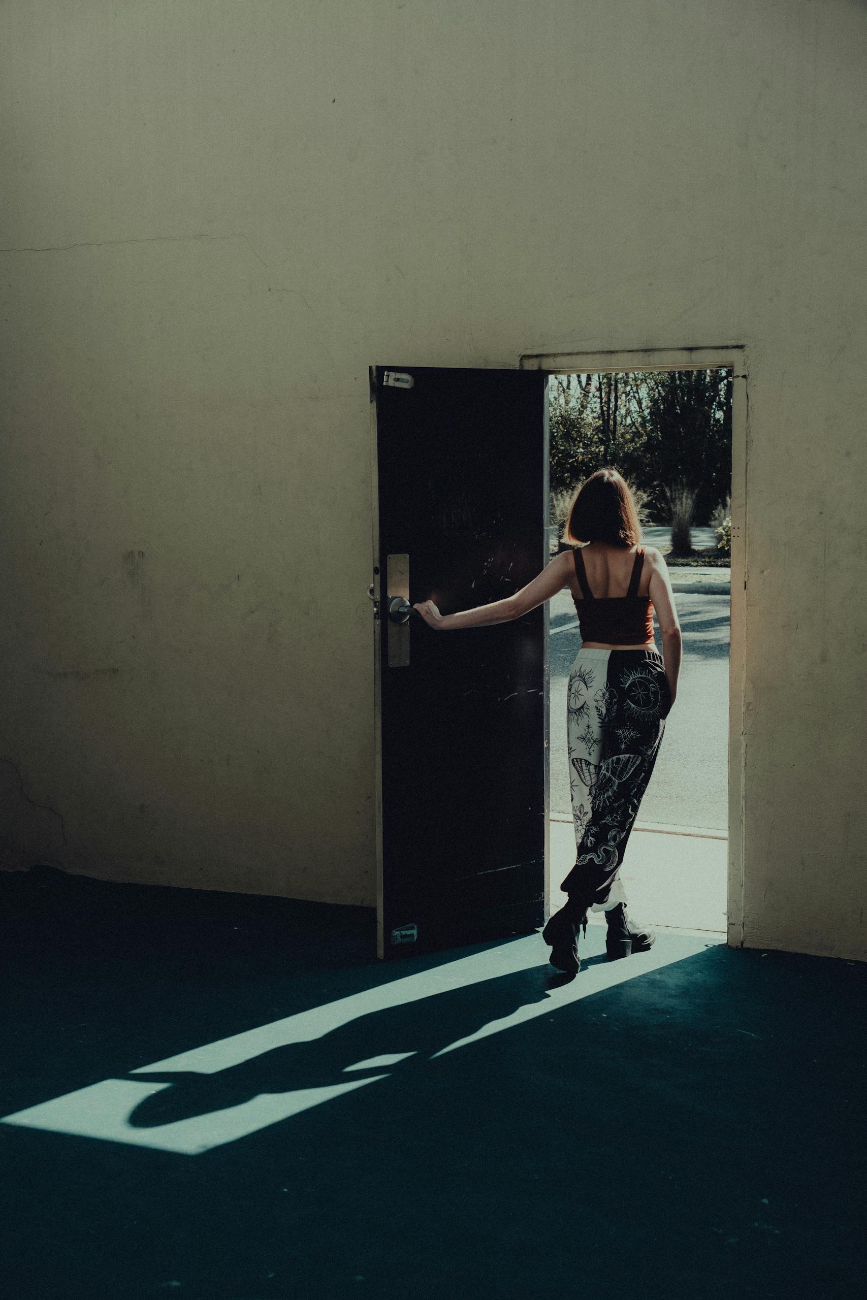 A woman stepping out into sunlight through an open doorway, casting a shadow.